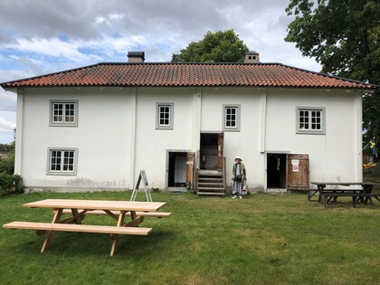 Vit tvåvåningsbyggnad med rött tegeltak, picknickbord på gräsmatta och en person i traditionella kläder.