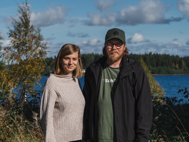 En kvinna och en man står utomhus vid en sjö med skog i bakgrunden under en solig dag.