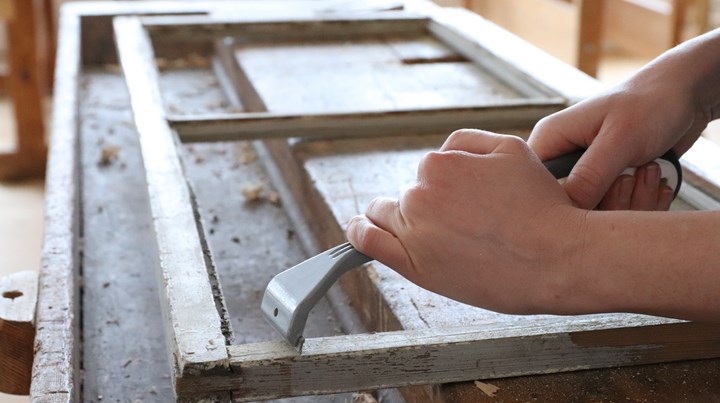 En person skrapar färg från en gammal träram med ett verktyg på en arbetsbänk.