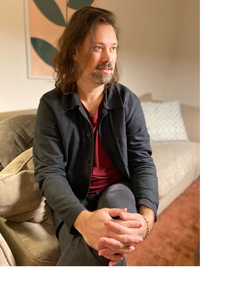Man med långt hår sitter på en soffa, händerna knäppta, tittar åt sidan i ett vardagsrum.