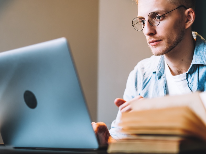 Ung man med glasögon studerar vid en bärbar dator med öppna böcker på ett bord.