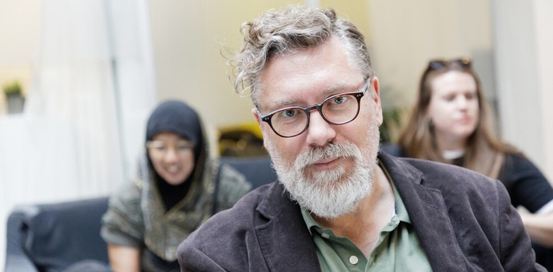 Man med glasögon och grått skägg sitter i fokus, två kvinnor i bakgrunden, inomhusmiljö.