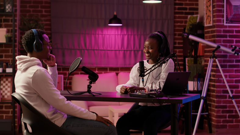 Två personer spelar in en podcast i en studio med mikrofoner och dator, omgivna av lila belysning.