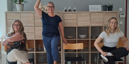 Tre kvinnor poserar lekfullt på och bredvid stolar i ett ljust kontorsrum med hyllor i bakgrunden.