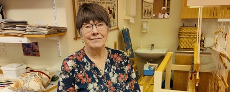 Äldre kvinna med glasögon står vid ett bord med färgglada tygstycken i en hantverksverkstad.