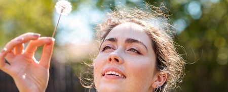 Kvinna i blå jacka håller en maskros och tittar upp mot den utomhus en solig dag.