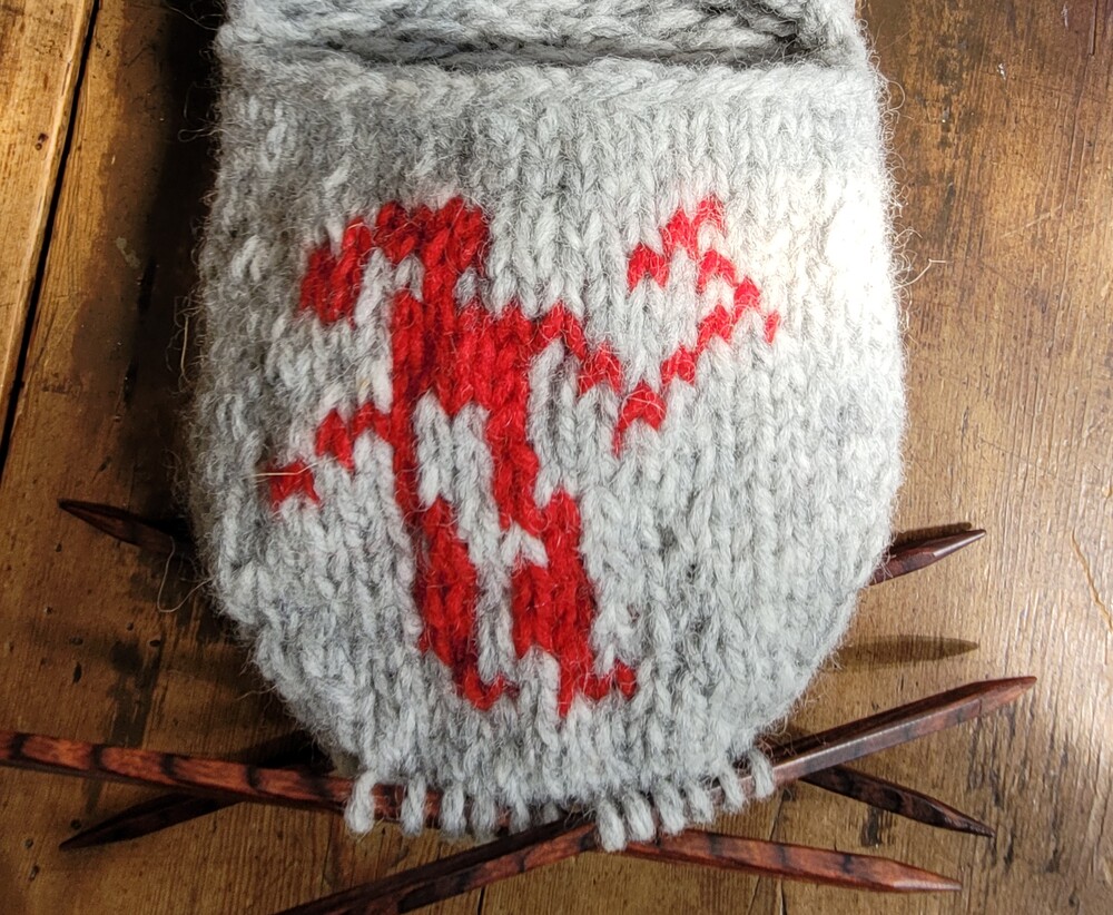 Stickat grått fodral med röd mönsterfigur, liggande på träbord med strumpstickor.