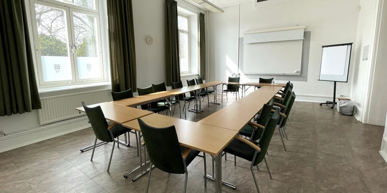 Ljust konferensrum med U-formad bordsuppställning, svarta stolar, stora fönster och whiteboard.