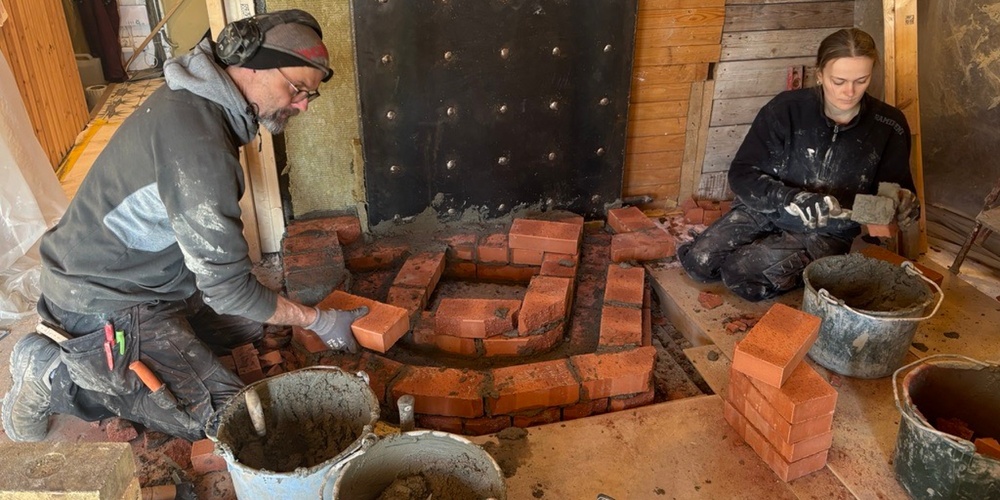 Två personer bygger en mur av tegelstenar inomhus, omgivna av verktyg och murbrukshinkar.
