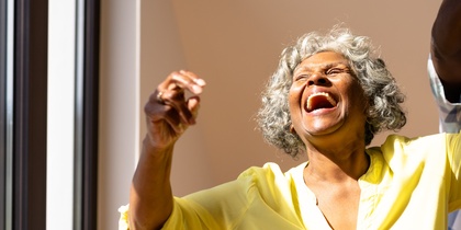 Äldre kvinna i gul skjorta skrattar och dansar med en man i ett soligt rum.