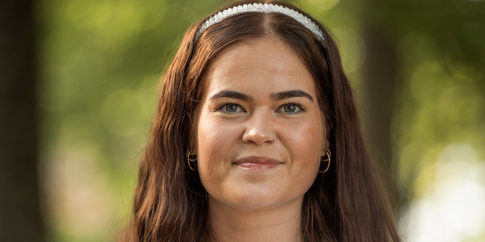 Ung kvinna med långt brunt hår och vit blus står utomhus med suddig grön bakgrund.