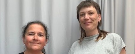 Två personer står bredvid varandra framför ett grått draperi och ler mot kameran.