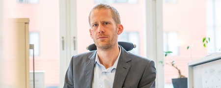 Man i kostym sitter på ett kontor framför ett fönster, tittar mot kameran.