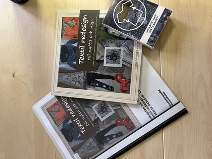En studieplan, en bok och en kortlek om Textil ReDesign som ligger på ett ljust träbord.
