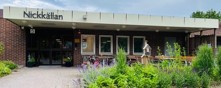 Entré till byggnad med skylten 'Nickkällan', omgiven av grönska och blommor under en molnig himmel.