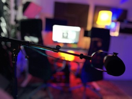 Närbild på en mikrofon i ett färgglatt, suddigt inspelningsstudio med dator i bakgrunden.