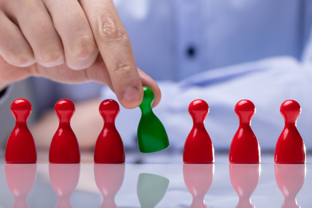 En hand lyfter en grön spelpjäs bland flera röda pjäser på ett blankt bord.