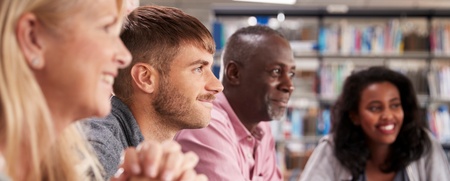 Fyra vuxna personer sitter vid ett bord i ett bibliotek och arbetar tillsammans med datorer och anteckningar.