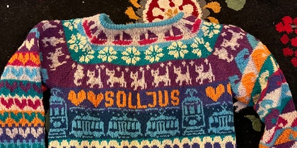 Färgglad stickad tröja med mönster, text och symboler på en blommig matta.
