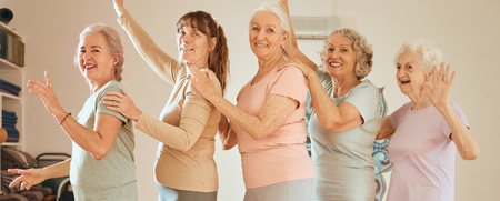 Fem äldre kvinnor gör yoga eller dansar tillsammans i ett ljust träningsrum med trägolv.