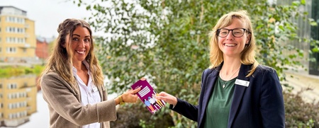 Två personer utbyter en broschyr utomhus vid en vattendrag och grönska.