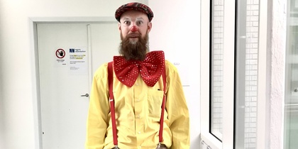 Person i clownliknande klädsel med stor röd fluga, gula skjorta, hängslen och ukulele i en ljus korridor.