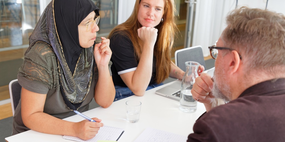 Tre personer sitter vid ett bord och diskuterar, med anteckningsblock och glas vatten framför sig.