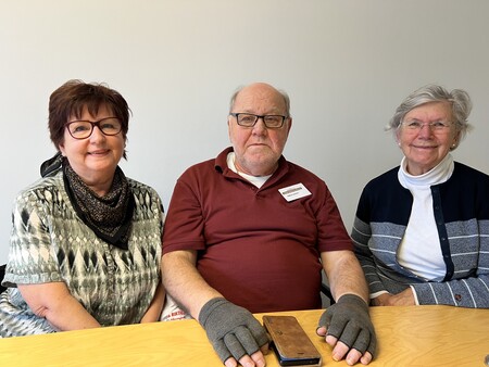 Tre äldre personer sitter vid ett bord, två kvinnor och en man, alla ler mot kameran.