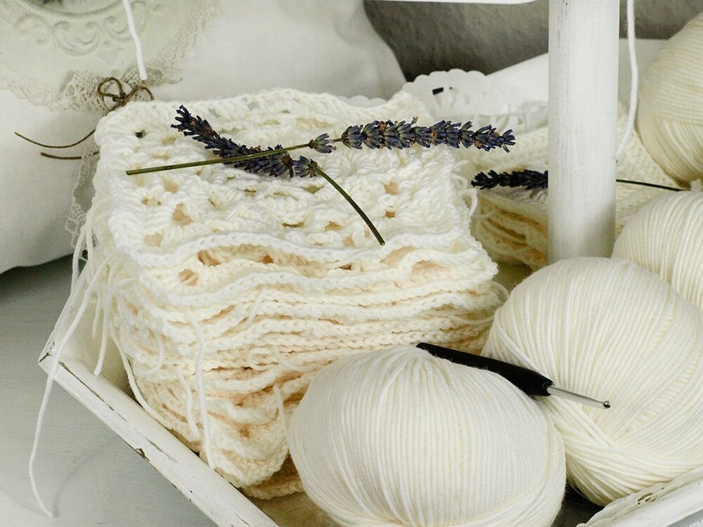 Vita virkade rutor, garnnystan och en virknål på en bricka med lavendelkvistar.