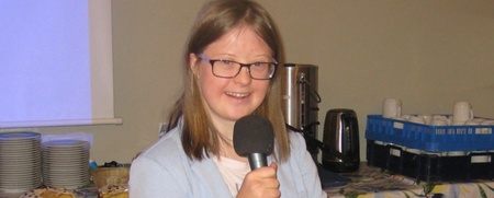 Ung kvinna med glasögon håller en mikrofon och ler, står framför ett bord med porslin och fika.