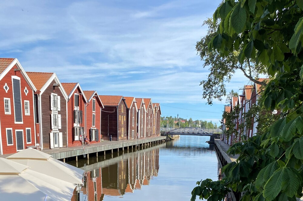 Röda trähus vid en kanal med spegelblankt vatten och grönska i förgrunden, blå himmel ovanför.
