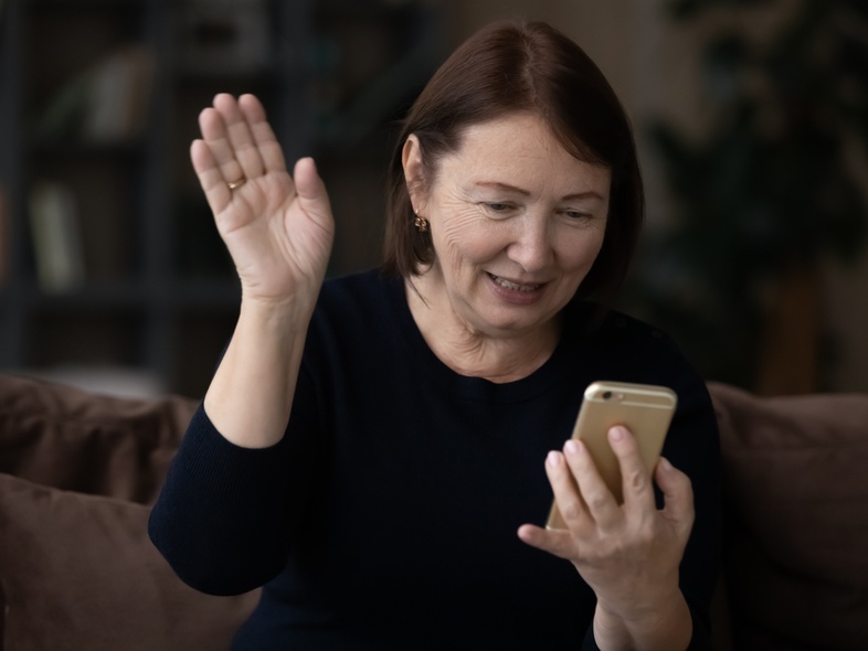 Äldreperson med svart tröja och brunt hår vinkar och ler mot en mobiltelefon, sitter i soffa inomhus med mörk bakgrund