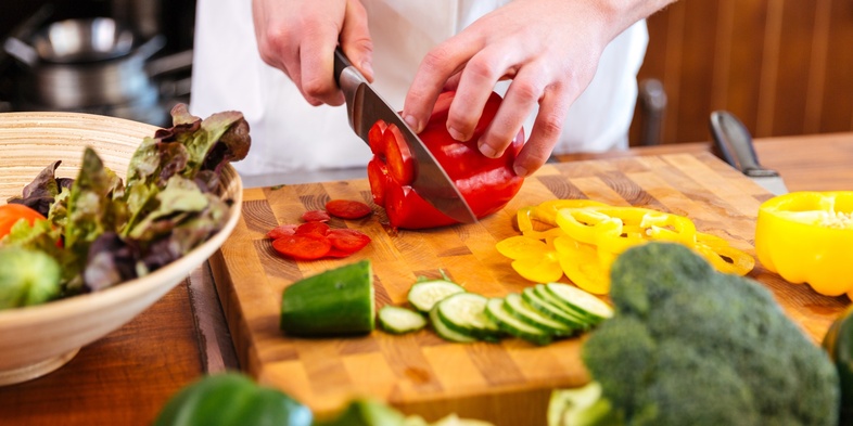 En person skär röda paprikor på en skärbräda omgiven av olika grönsaker i ett kök.