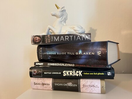 En stapel böcker med en vit enhörningsfigur ovanpå, placerade på ett vitt bord mot en ljus vägg.