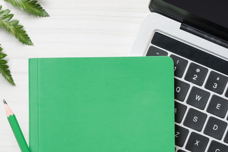 Grön anteckningsbok och penna på en bärbar dator, med växt och ormbunksblad på ett vitt skrivbord.