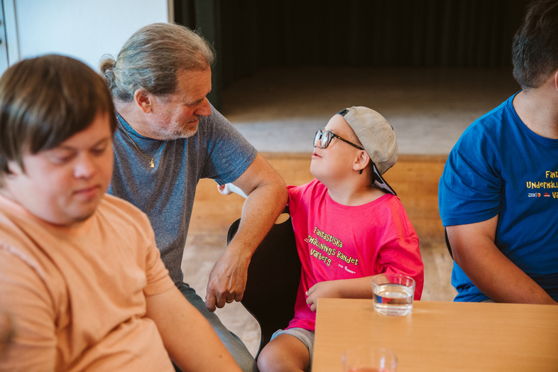 En äldre man och ett barn med keps och glasögon samtalar vid ett bord med fler personer.