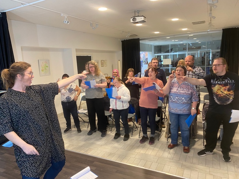 En grupp människor står i ett rum och sjunger, ledda av en kvinna som dirigerar dem.