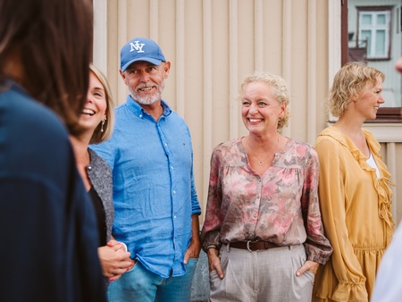 En grupp vuxna står utomhus framför en beige vägg och samtalar.