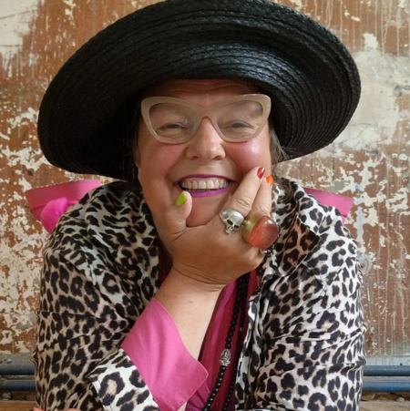 Leende person med stor svart hatt, leopardmönstrad jacka och färgglada accessoarer mot sliten vägg.