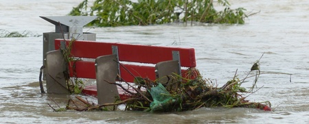 En röd parkbänk är delvis täckt av vatten och grenar efter en översvämning.