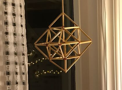 En geometrisk dekoration av sugrör hänger i ett fönster bredvid ett vitt gardin och ljusslingor syns i bakgrunden.