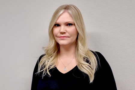 Kvinna med långt blont hår och svart tröja står mot en ljus vägg.