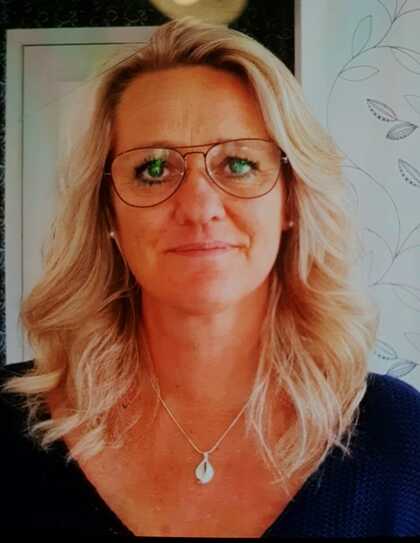 Kvinna med axellångt blont hår och halsband står framför en ljus bakgrund med bladmönster.