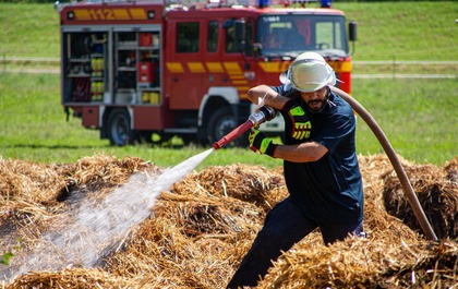 Brandman i hjälm släcker eld med vattenslang på ett fält, brandbil syns i bakgrunden.
