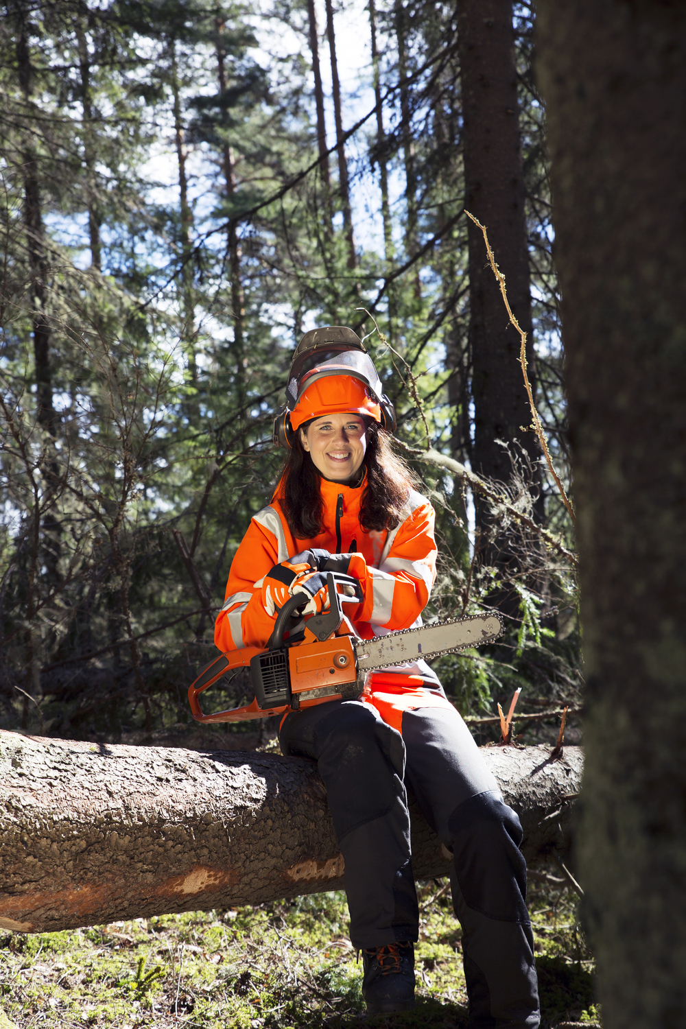 En kvinna i orange skyddskläder sitter på en fälld trädstam i skogen med en motorsåg i händerna.