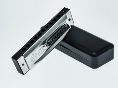 En munspel i metall och plast ligger lutad mot ett svart fodral på vit bakgrund.