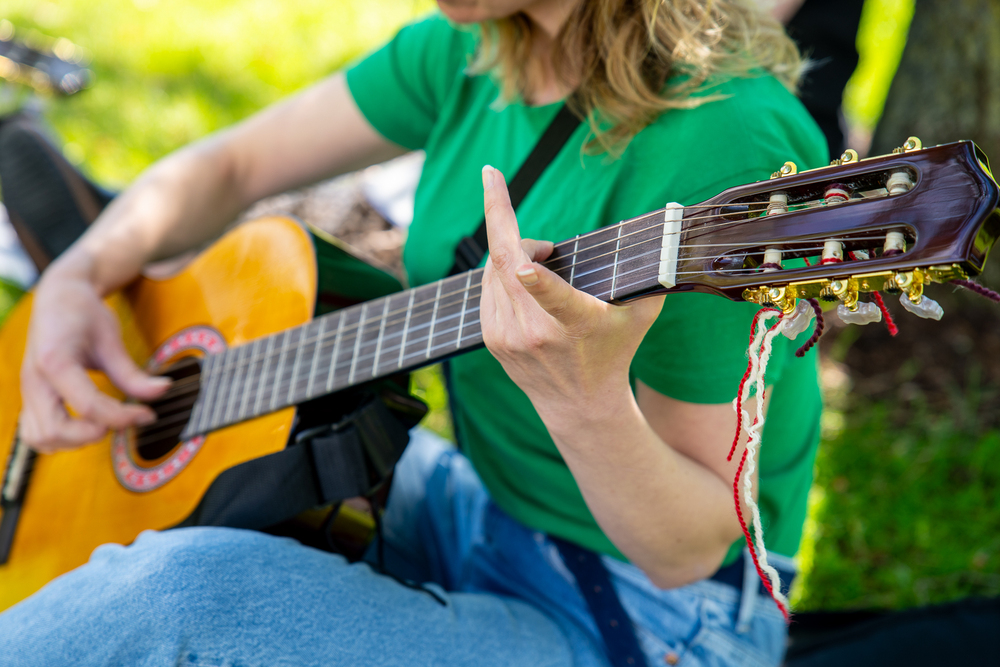 Person i grön tröja spelar akustisk gitarr utomhus, fokus på händer och gitarr.
