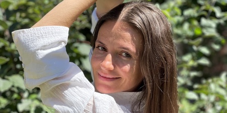 Kvinna med långt brunt hår poserar utomhus i solljus, omgiven av gröna växter.