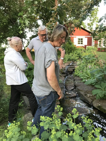 Tre vuxna personer står vid en liten bäck i en grön trädgård med ett rött hus i bakgrunden.