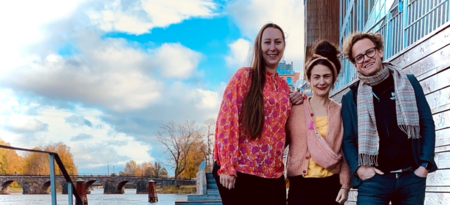 Tre personer står utomhus vid en flod med en bro och träd i bakgrunden, leende mot kameran.